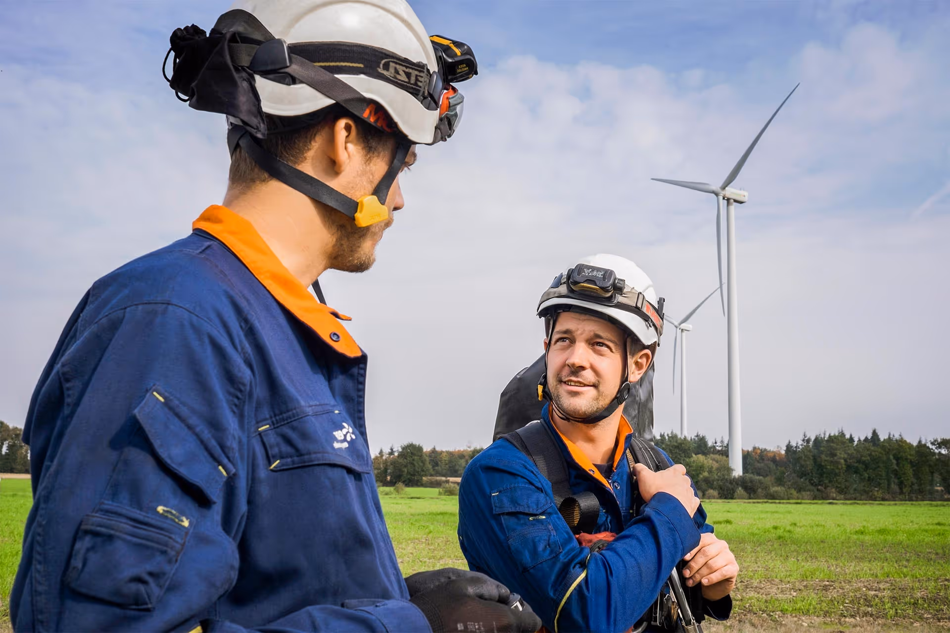 Two maintenance technicians wearing safety helmets and blue uniforms with orange collars standing in a green field with wind turbines in the background.