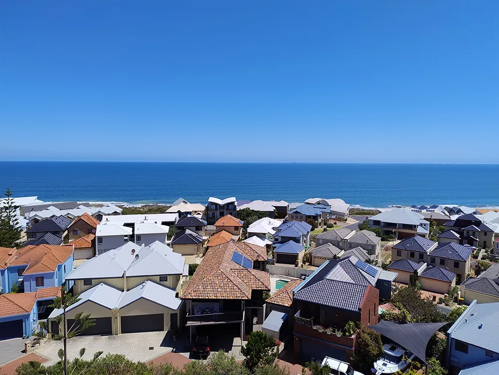 A coastal neighborhood with modern houses and tiled roofs overlooking a calm blue ocean under a clear sky.