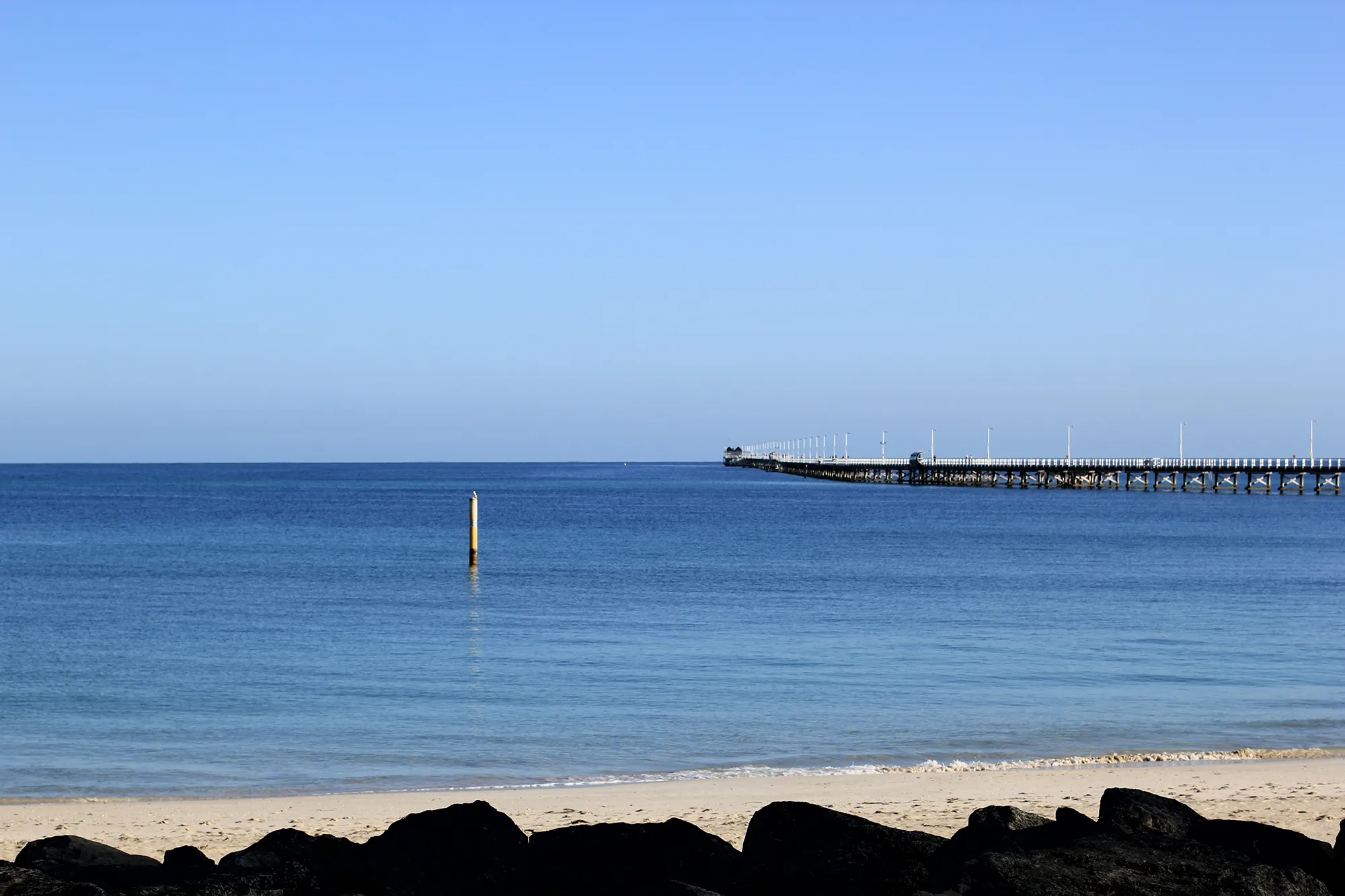 Calm blue ocean with a sandy beach, dark rocks in the foreground, and a long wooden pier extending into the water under a clear sky.