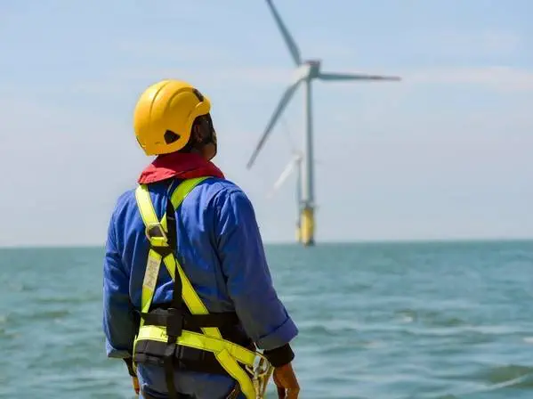Worker wearing safety harness and yellow helmet looking at offshore wind turbines over the sea.