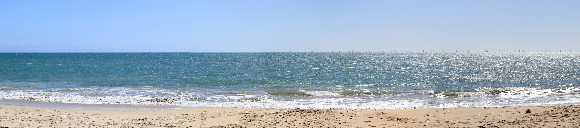 Sunny beach with light waves, sandy shore, and wind turbines visible on the horizon over the ocean.