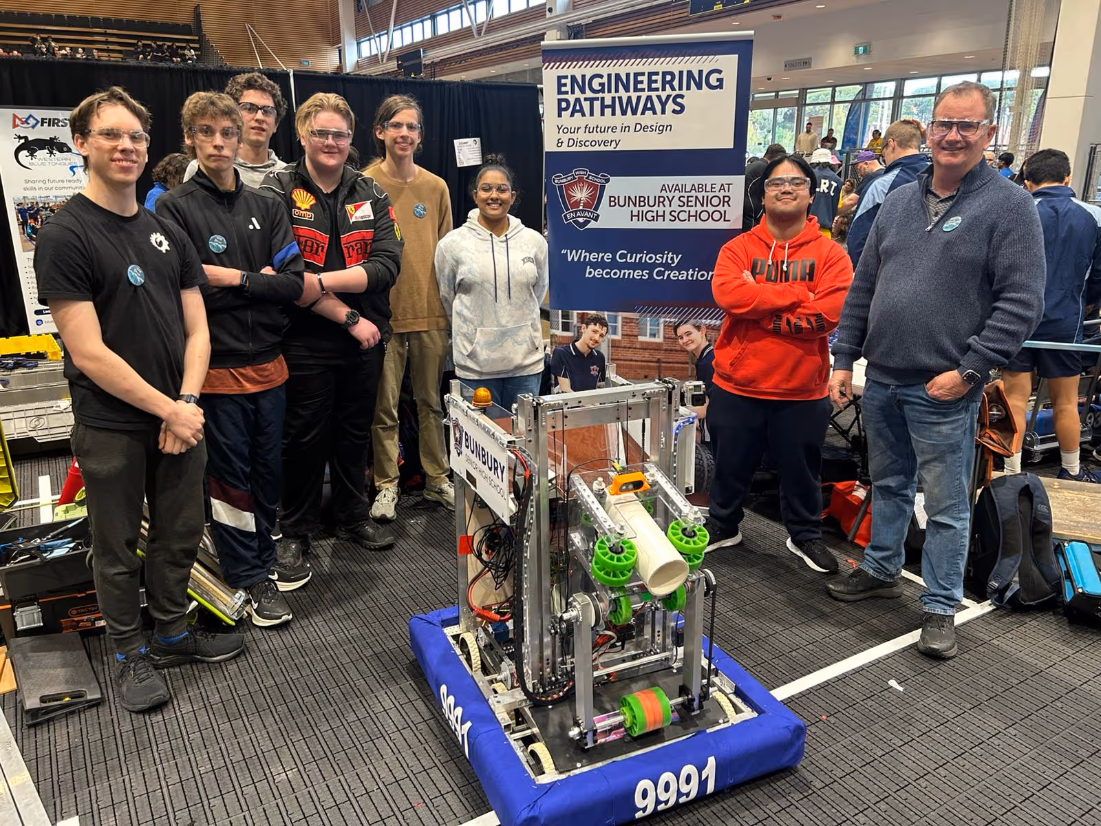Group of diverse students and a teacher standing behind a robotics project with a sign that reads Engineering Pathways at Bunbury Senior High School.