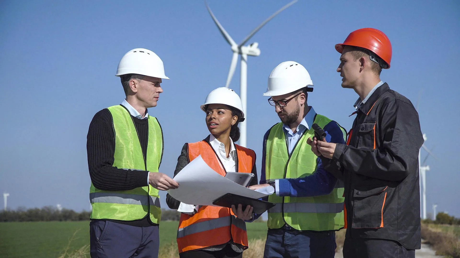 Four engineers wearing safety vests and helmets discussing plans outdoors near wind turbines.