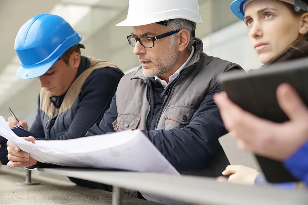 Three construction workers wearing helmets reviewing blueprints and taking notes on a site.