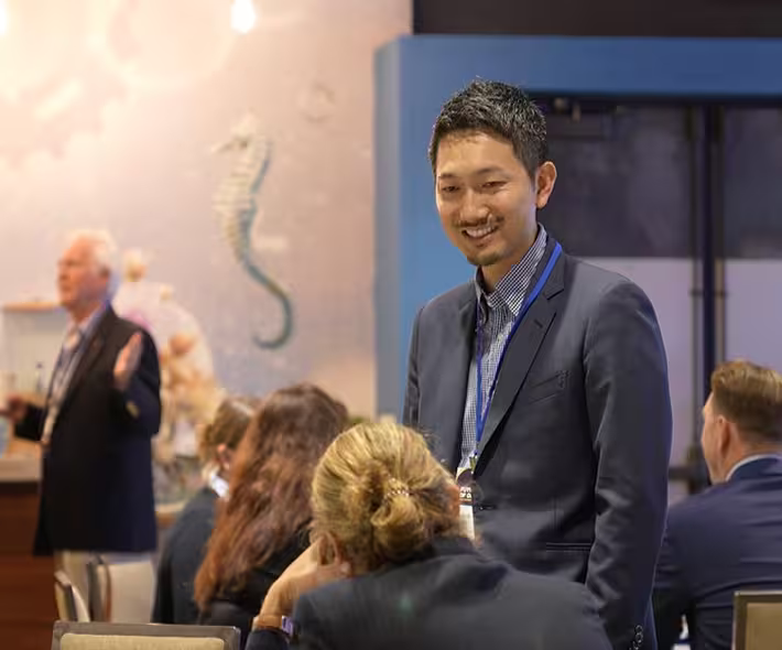 Smiling man in a suit and name badge conversing with seated people in a conference setting.