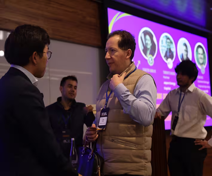 Four men engaged in conversation at an indoor event with a purple presentation screen in the background.