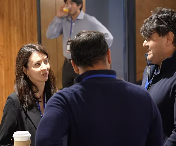 Three people engaged in conversation indoors, with one person in the background drinking from a glass.