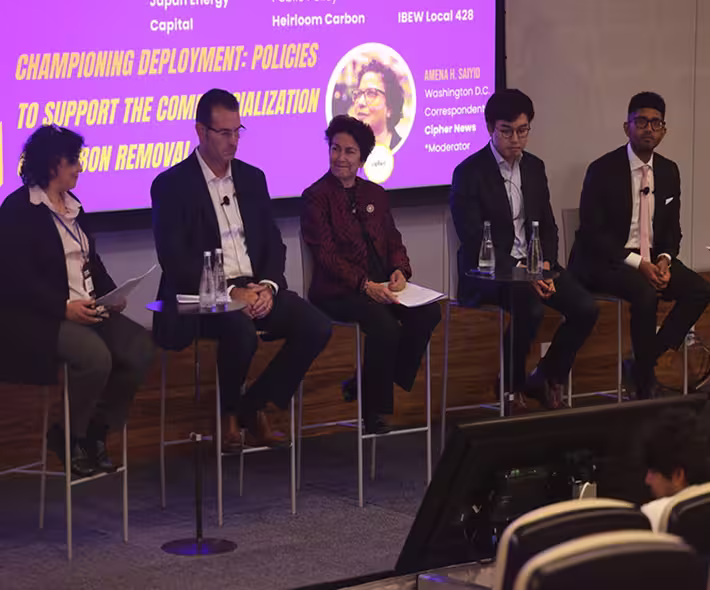 Five people seated on high stools on a stage panel discussion with a purple presentation slide behind them.