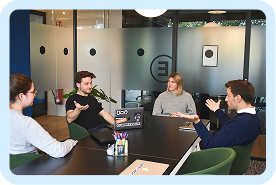 Four people engaged in a meeting around a conference table with laptops and notebooks.