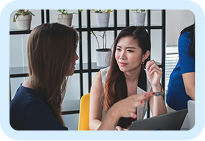 Two women engaged in a discussion while working on laptops in an office setting.