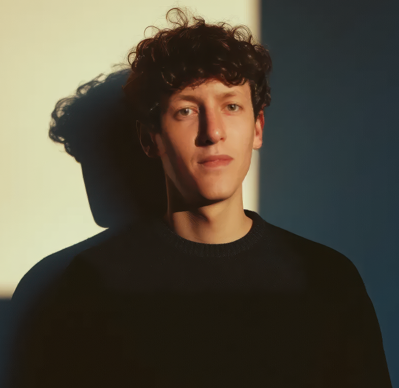 Young man with curly hair wearing a black sweater and looking at the camera with a shadow cast on the two-toned wall behind him.