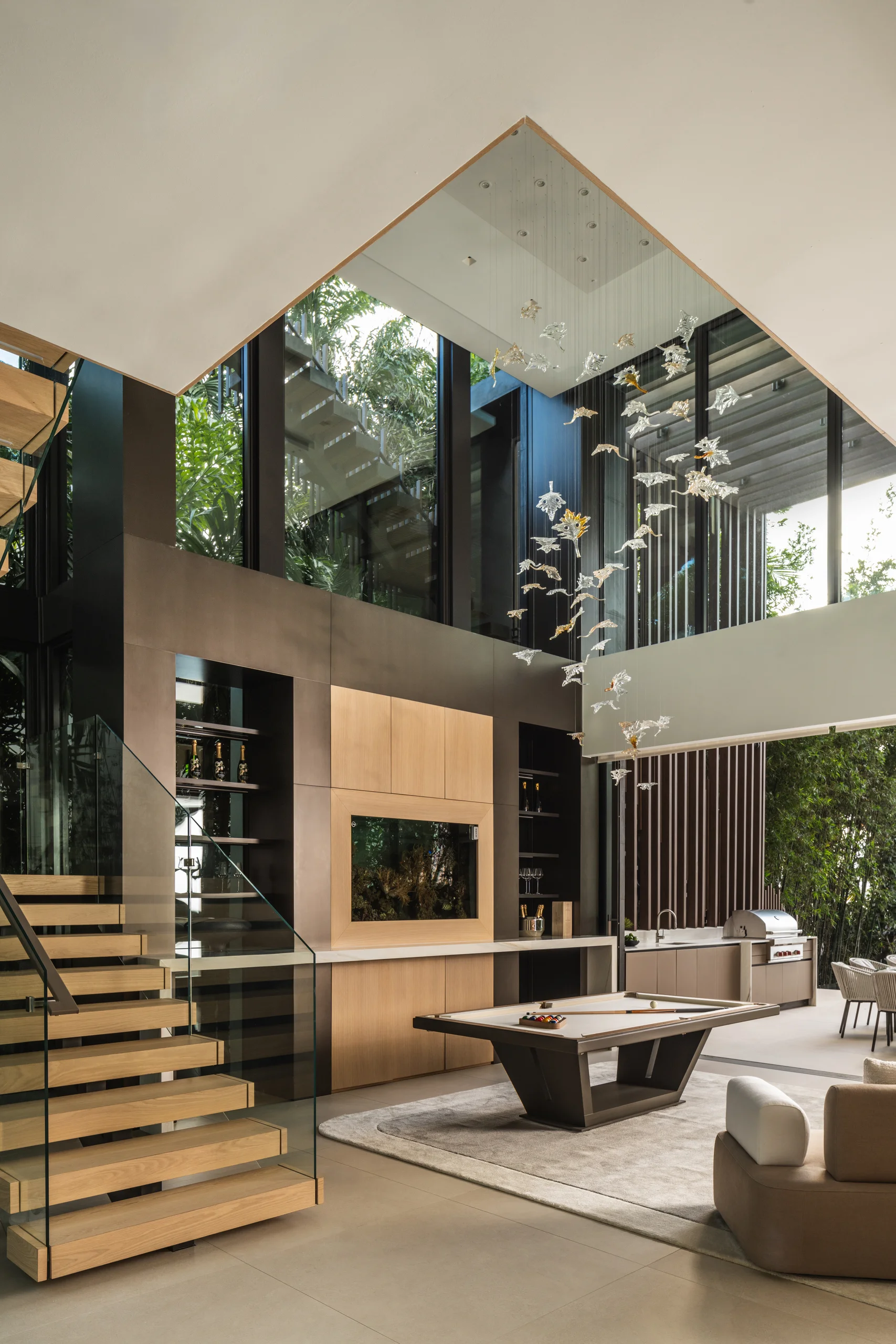 Double-height great room with a floating staircase, linear fireplace, and pool table opening to an outdoor kitchen, Miami's Rive Gauche project by Dunagan Design.