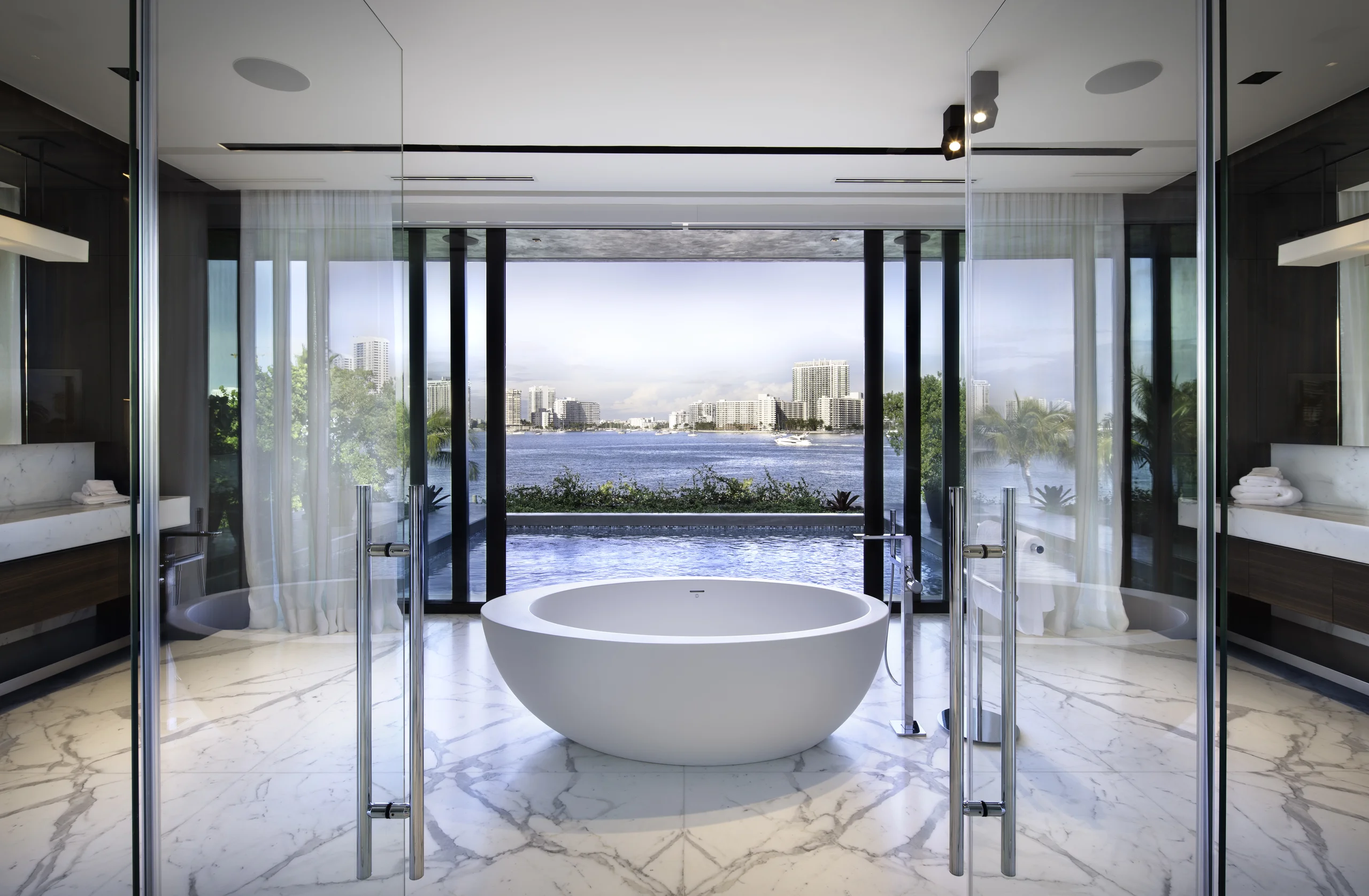 Primary bathroom with a freestanding oval tub facing a floor-to-ceiling glass wall and the Miami skyline, Star Island Waterfront project by Dunagan Design.