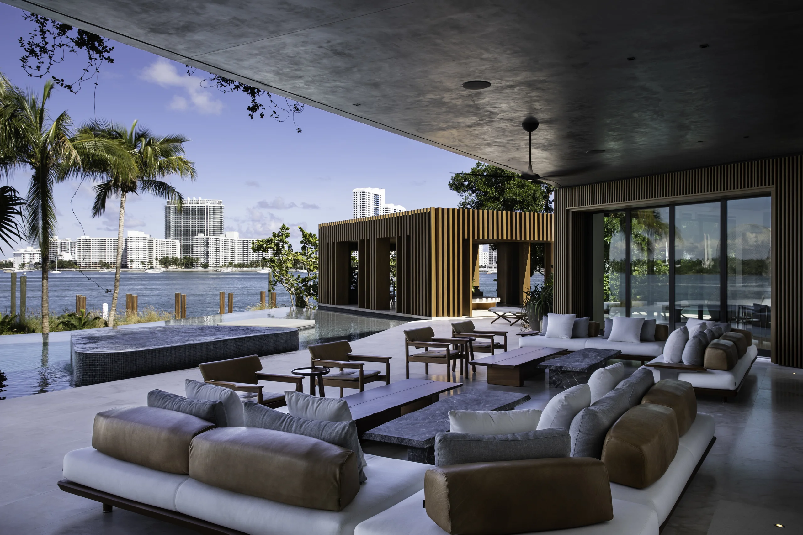 Covered outdoor loggia with sectional seating, an infinity pool and spa, and a wood-slat cabana against the Miami skyline, Star Island Waterfront project by Dunagan Design.