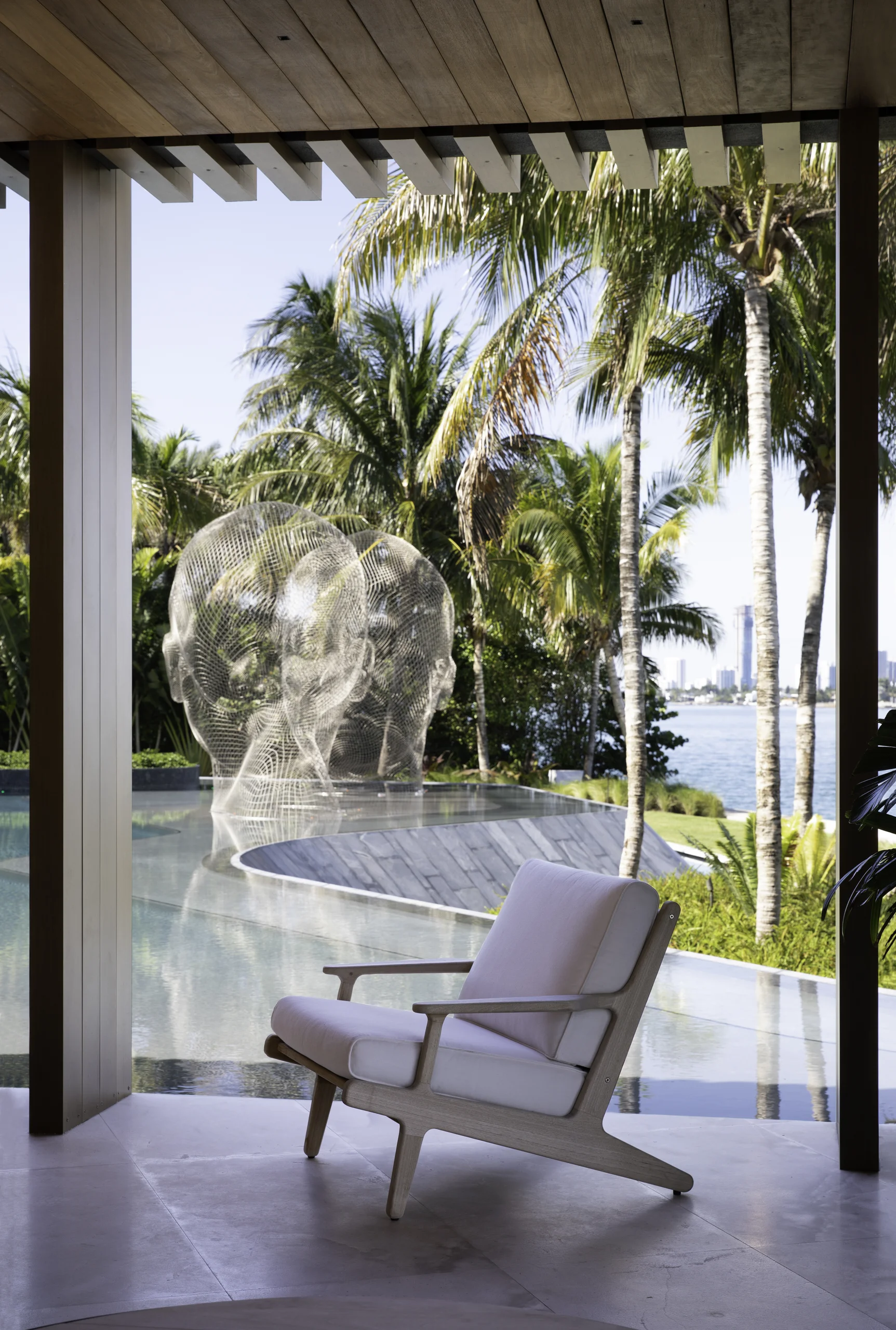 Wood-framed lounge chair on a shaded terrace overlooking the infinity pool, where Jaume Plensa's wire-mesh head sculptures rise among palm trees with the bay beyond, Star Island Waterfront project by Dunagan Design.