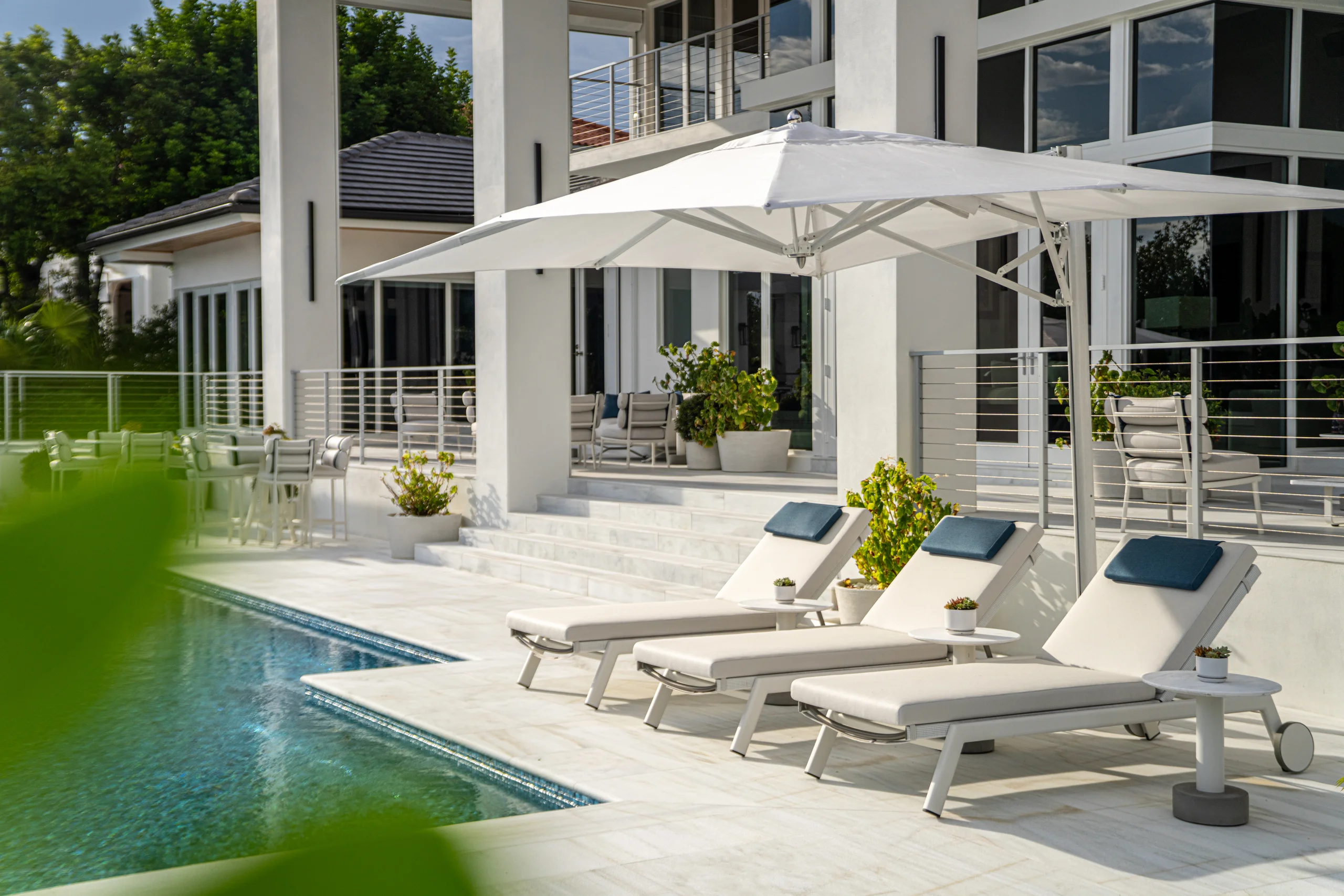 Pool terrace with cushioned chaise lounges, a cantilevered umbrella, and covered outdoor seating beyond, Coco Plum Revival project by Dunagan Design.