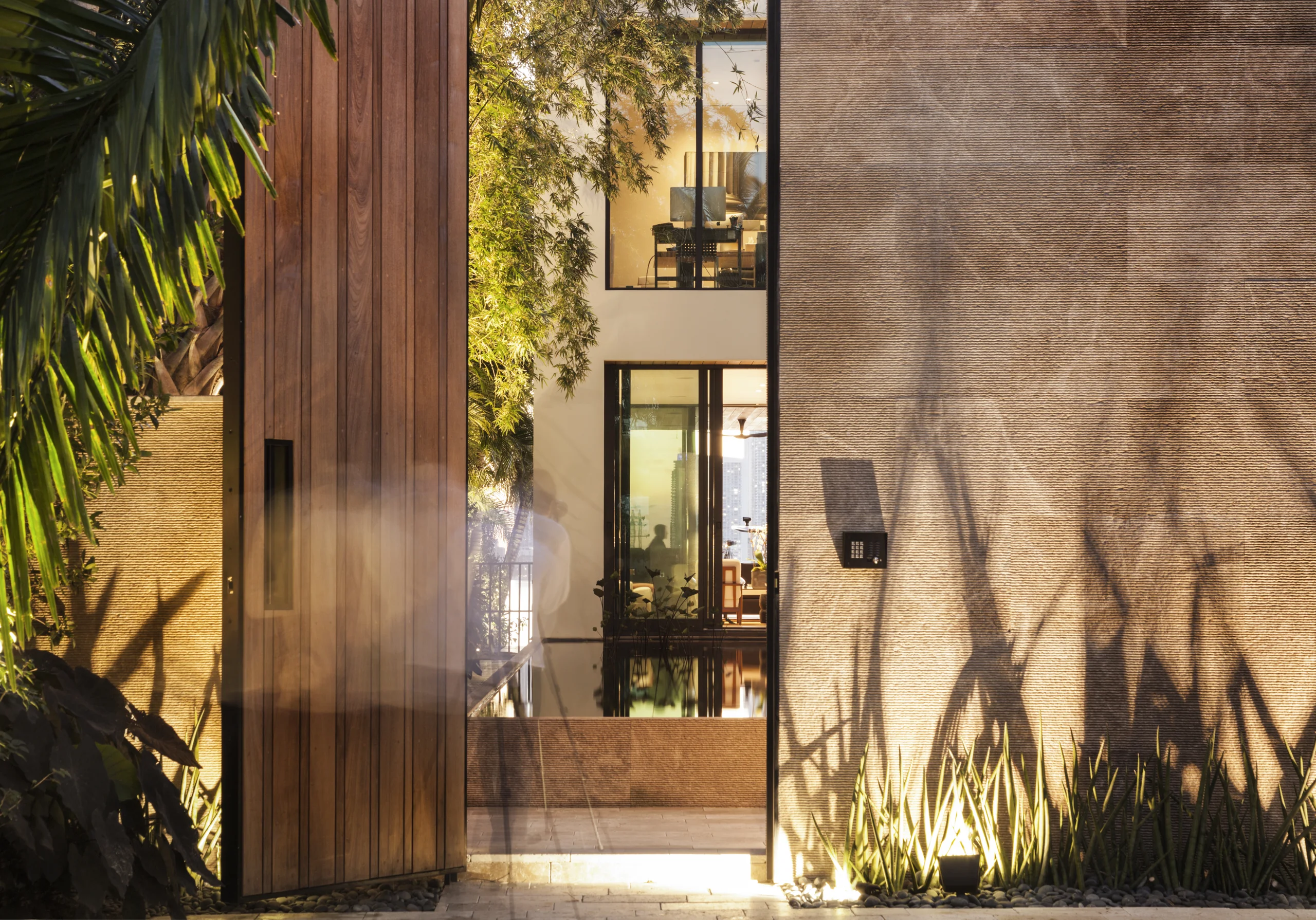 Wide-plank wood pivot entry door opening to a reflecting pool and glowing interior beyond, Bayside Modern project by Dunagan Design.