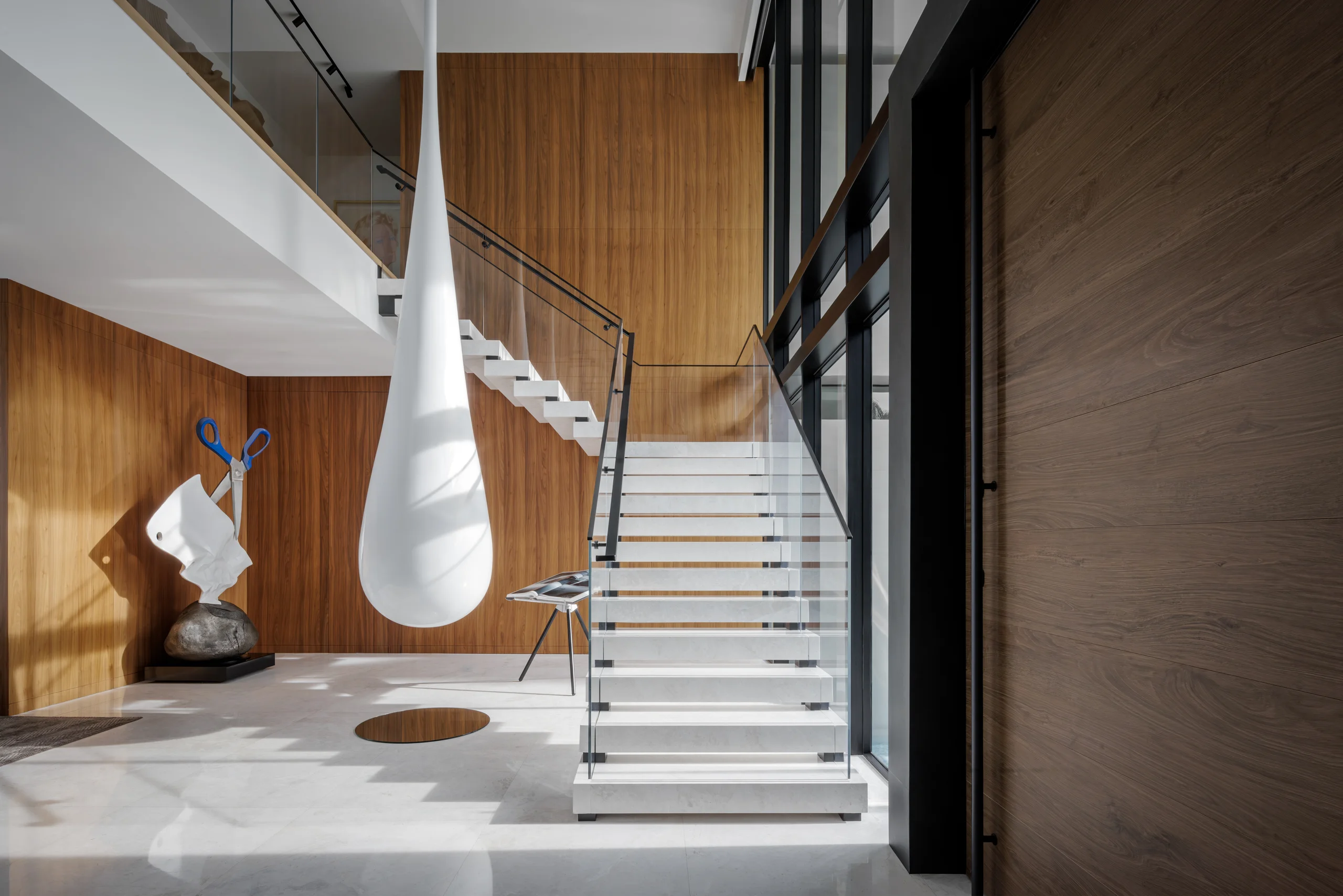 Double-height entry with a floating stone staircase, oversized white teardrop pendant, walnut-paneled walls, and a sculptural scissors artwork beside a walnut pivot door, San Marino Gallery project by Dunagan Design.