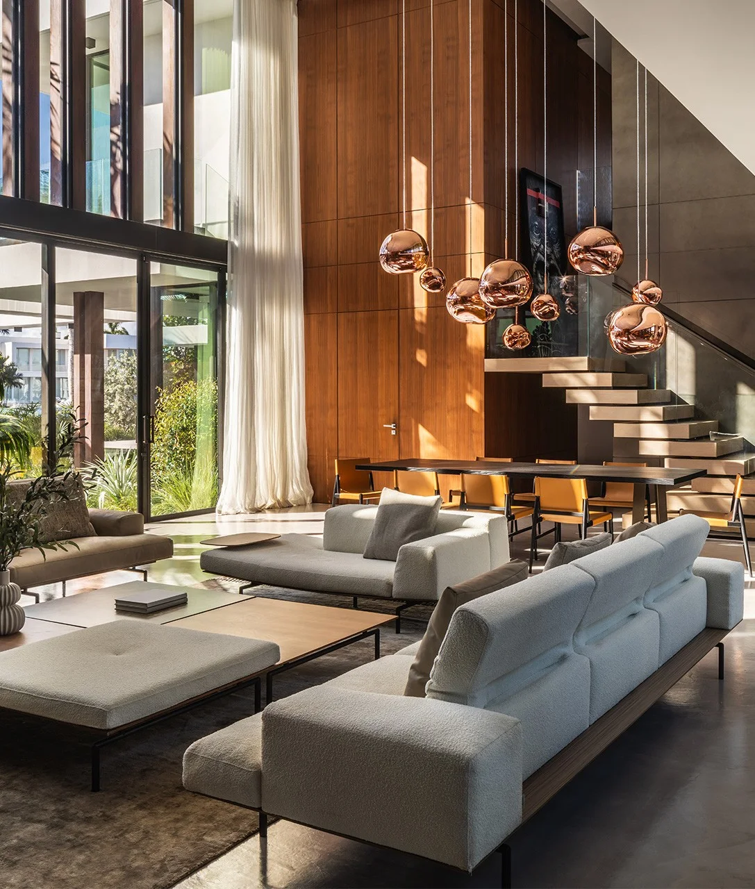 Double-height living and dining area with copper globe pendants, walnut-paneled walls, white bouclé sofas, and a floating stair beside floor-to-ceiling glass, Spanish River Modern project by Dunagan Design