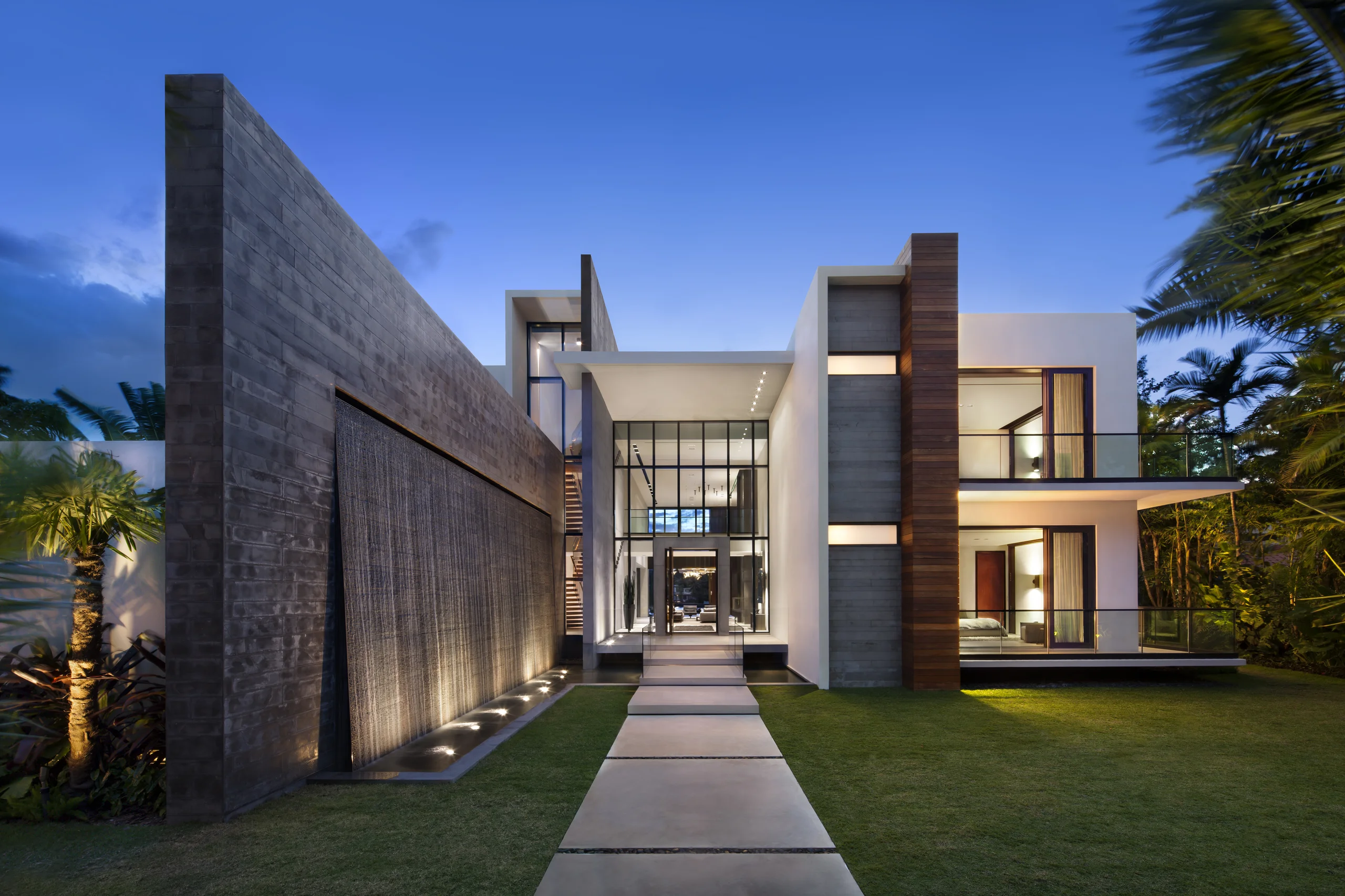 Twilight exterior with a stone water-wall feature, glass entry pavilion, and a floating stepping-stone walkway, DiLido Modern project by Dunagan Design. Entry detail of an oversized walnut pivot door beside a stone water-wall feature, DiLido Modern project by Dunagan Design. Glass entry pavilion at night opening to a fireplace lounge beyond, DiLido Modern project by Dunagan Design. Gallery entry with a large-scale abstract painting and a geometric brass mirror, DiLido Modern project by Dunagan Design. Living and dining area with a sculptural gold ring pendant cluster and a mirrored dark cabinetry wall, DiLido Modern project by Dunagan Design. Great room with a ring chandelier and a stone-clad fireplace column, open to the dining area beyond, DiLido Modern project by Dunagan Design. Open living, kitchen, and dining area with a gold bamboo-style chandelier and a leather sectional, DiLido Modern project by Dunagan Design. Dining room at dusk beneath a dramatic brass pipe pendant fixture,