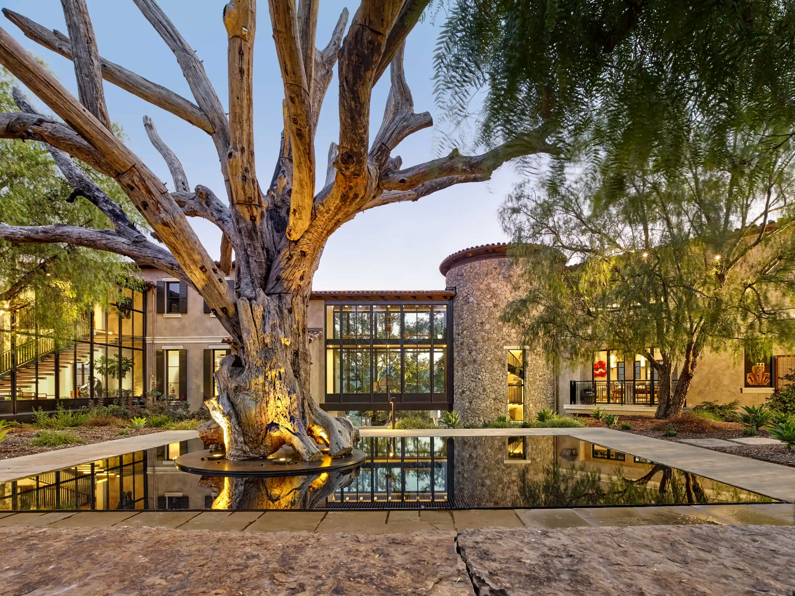 Courtyard at dusk with the weathered tree trunk uplit in the reflecting pool and warm light glowing from the steel-framed windows of the surrounding wings, Napa Valley Estate project by Dunagan Design.