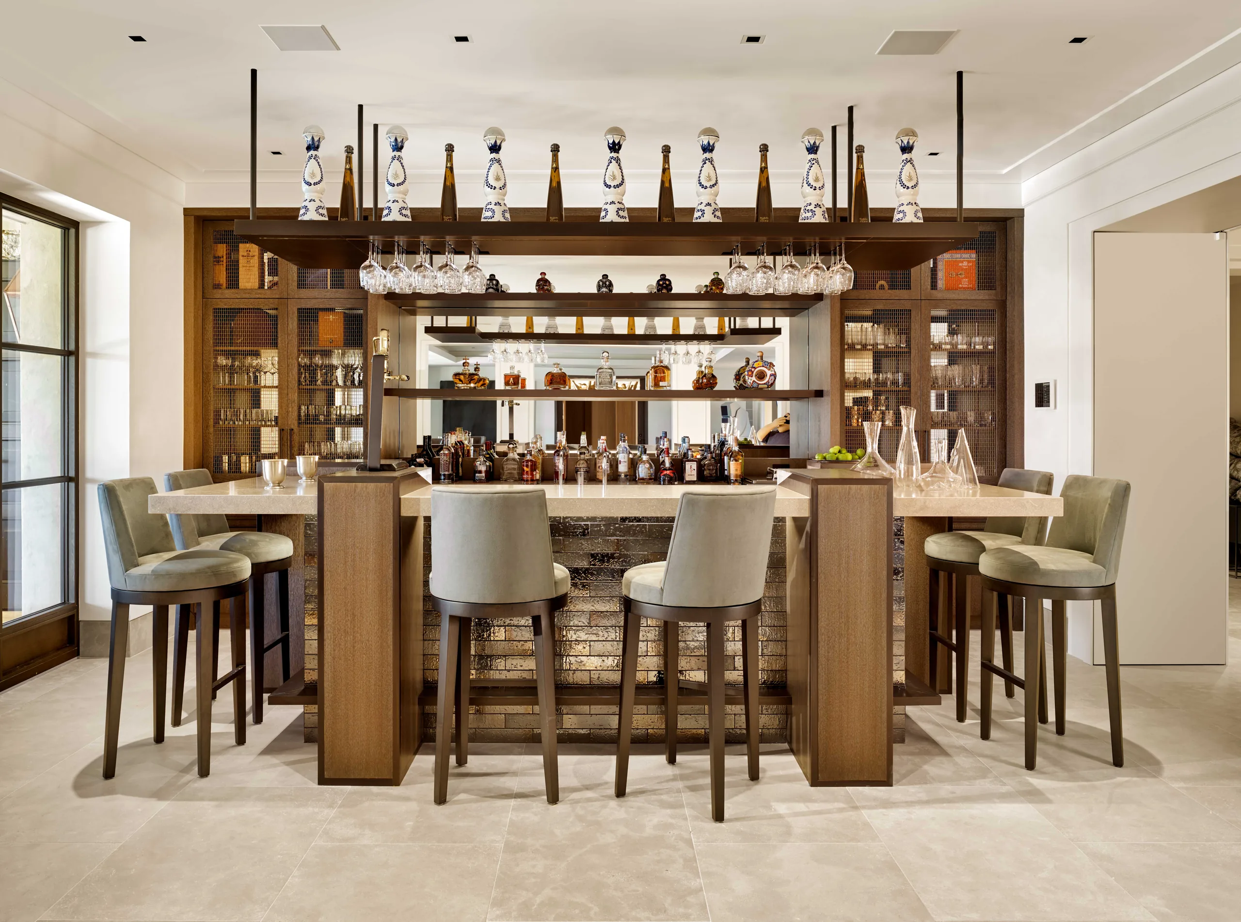 Home bar with a stone-topped counter, upholstered swivel stools, oak shelving, and a display of decorative tequila bottles above hanging stemware, Napa Valley Estate project by Dunagan Design.