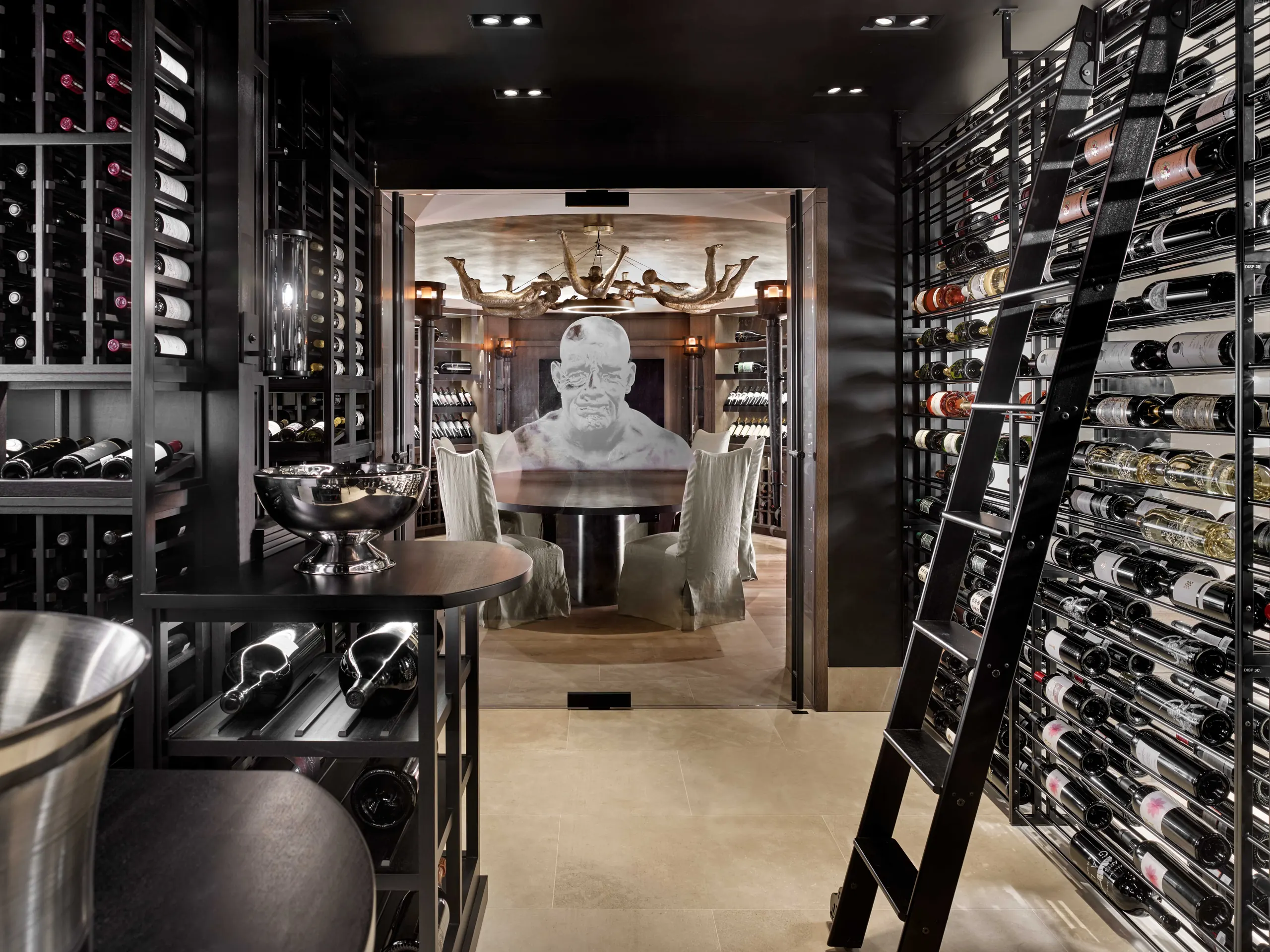 Wine cellar with black steel racking, a library ladder, and a tasting table beneath a sculptural chandelier, with a large white head sculpture beyond, Napa Valley Estate project by Dunagan Design.