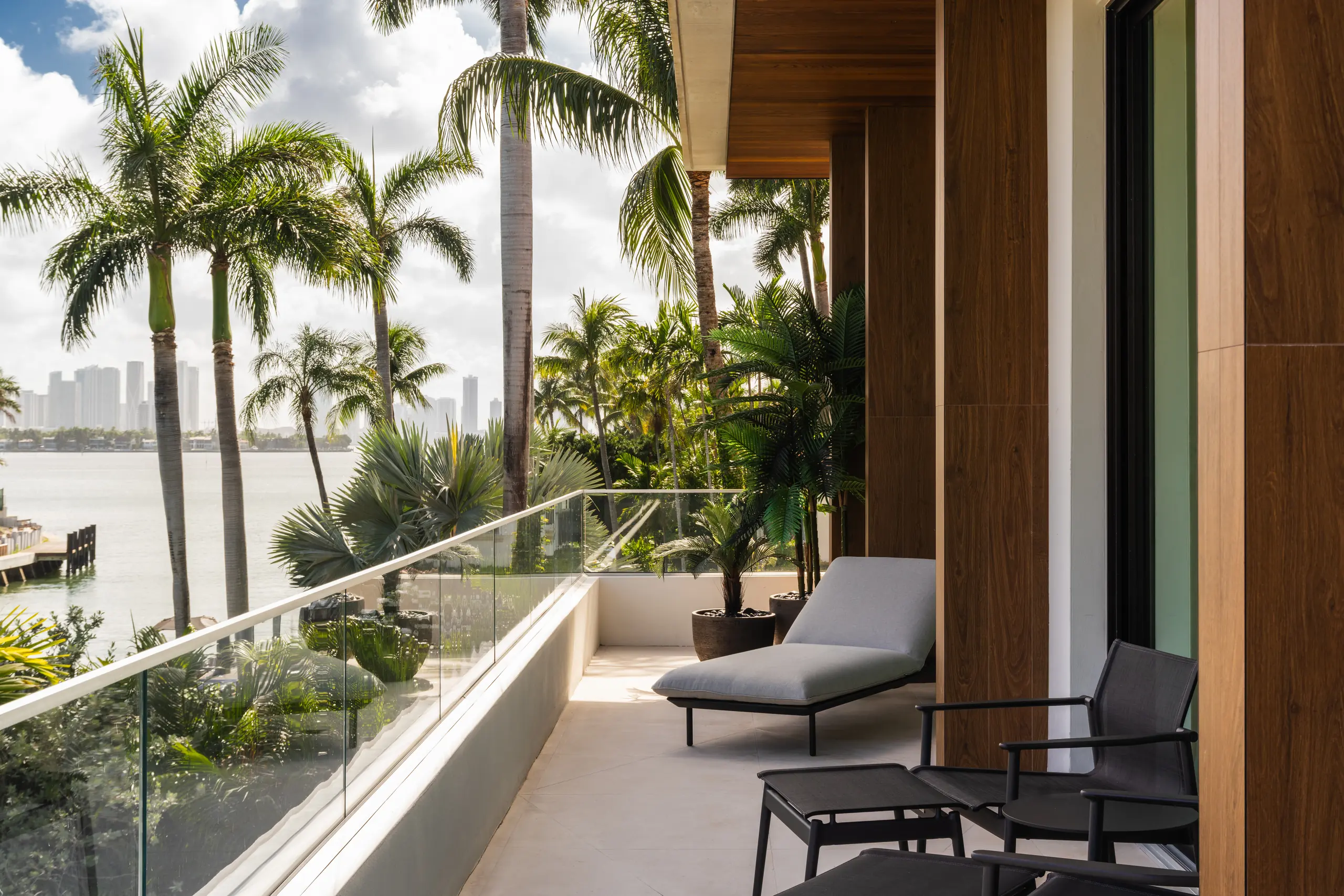Second-floor balcony with a glass railing and lounge seating overlooking palms and the bay, with the Miami skyline in the distance, Sunset Island House project by Dunagan Design.