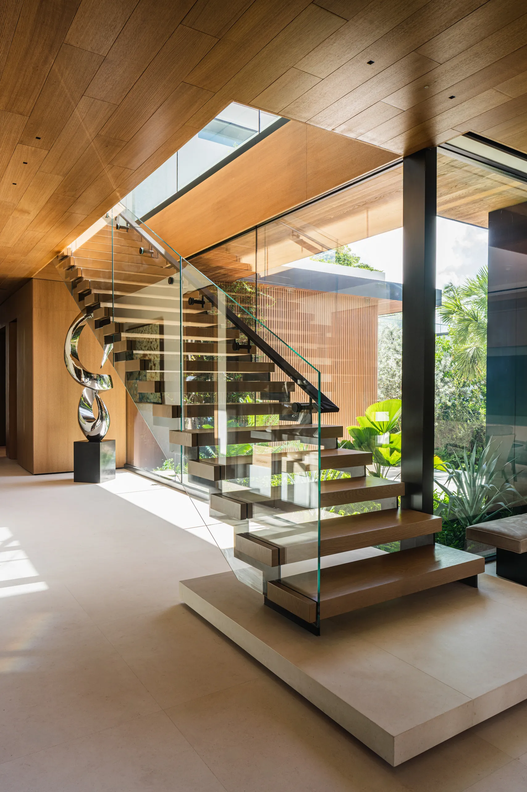 Floating oak staircase with a glass railing and a polished chrome sculpture at its base, set beneath a warm wood ceiling, Sunset Island House project by Dunagan Design.