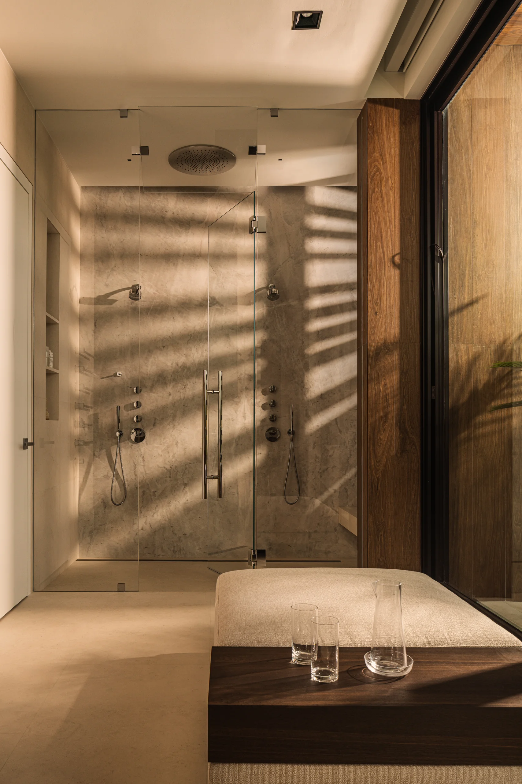 Walk-in shower clad in stone with a built-in bench, rain shower head and glass enclosure, with striped light cast through slatted screens, Sunset Island House project by Dunagan Design.