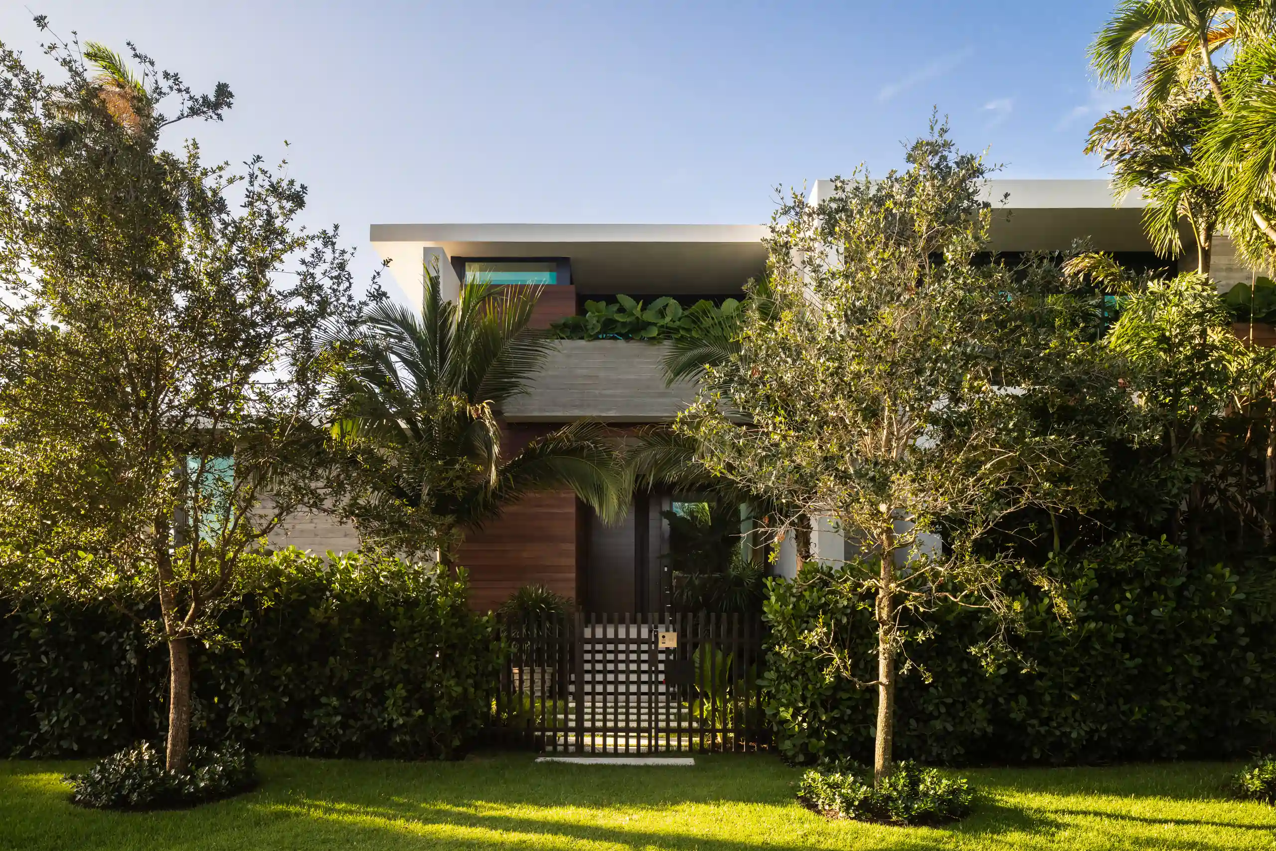 Street-facing entrance of a modern residence with a wood-clad entry, a slatted gate, mature trees and lush landscaping, Miami Beach Jewel project by Dunagan Design.