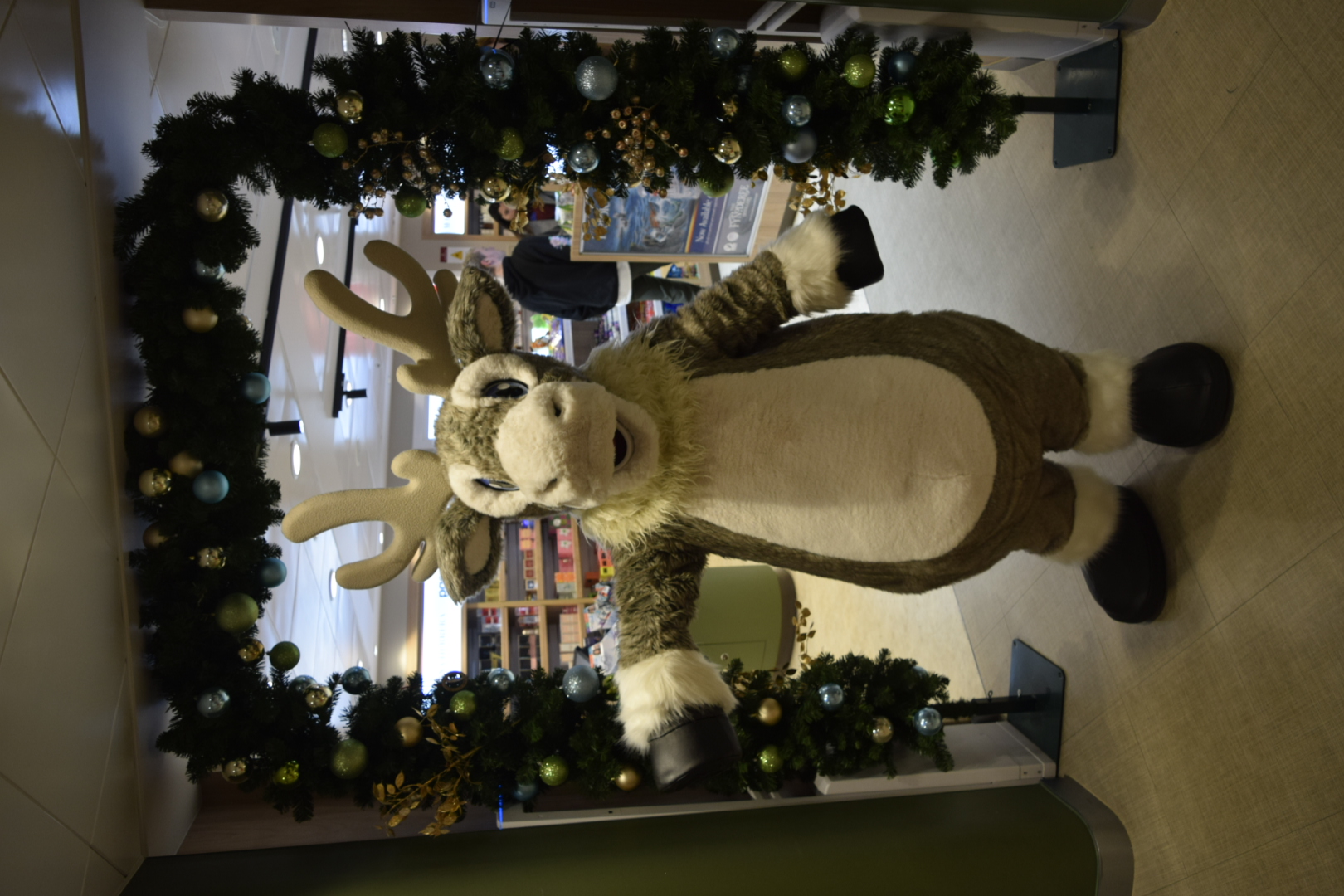 Arlo the reindeer is stood underneath a festive arch at the entrance of a store. He is waving at the camera.