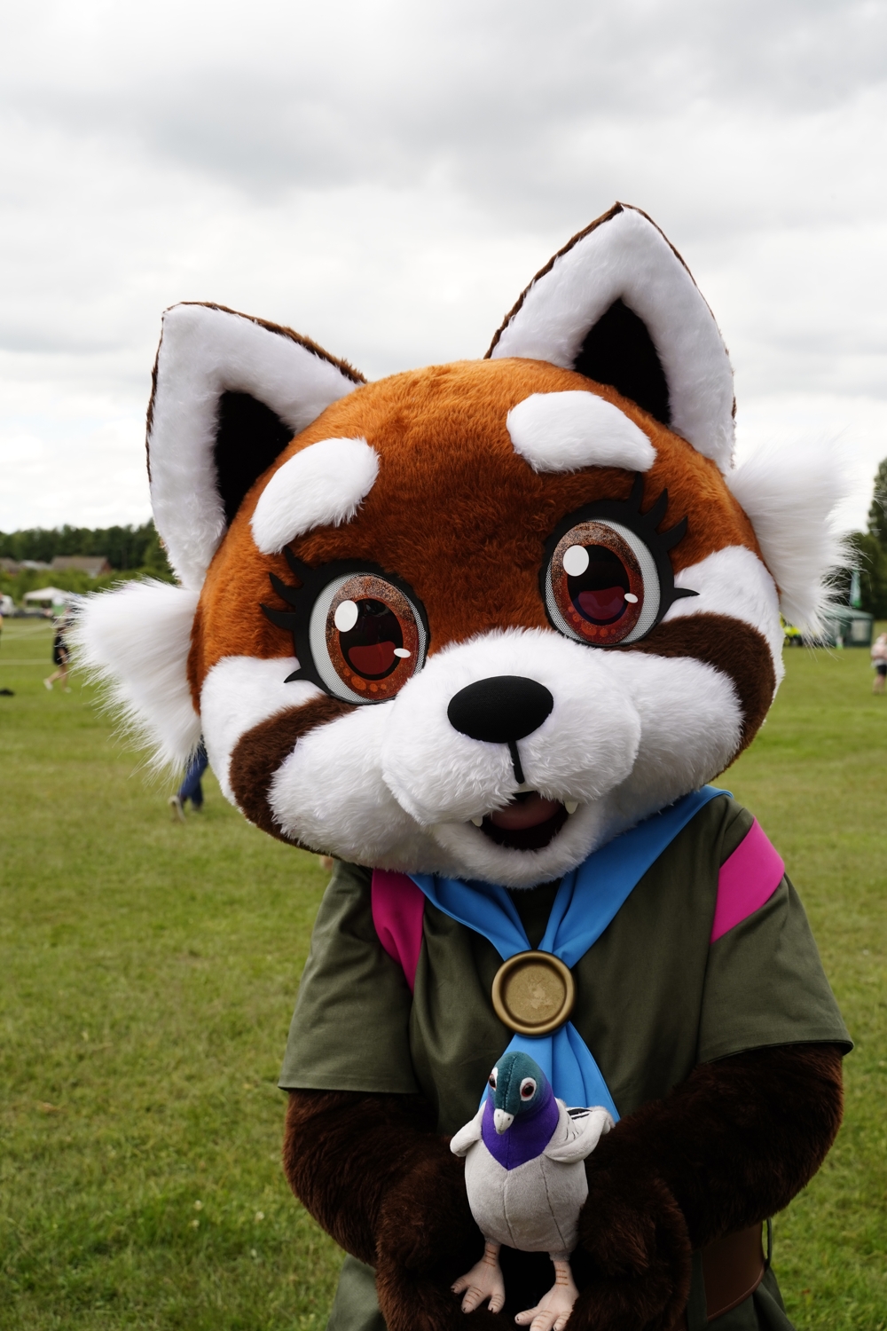 Zoomed in photo of Maple holding her toy pigeon in her paws. A festival ground can be seen behind her