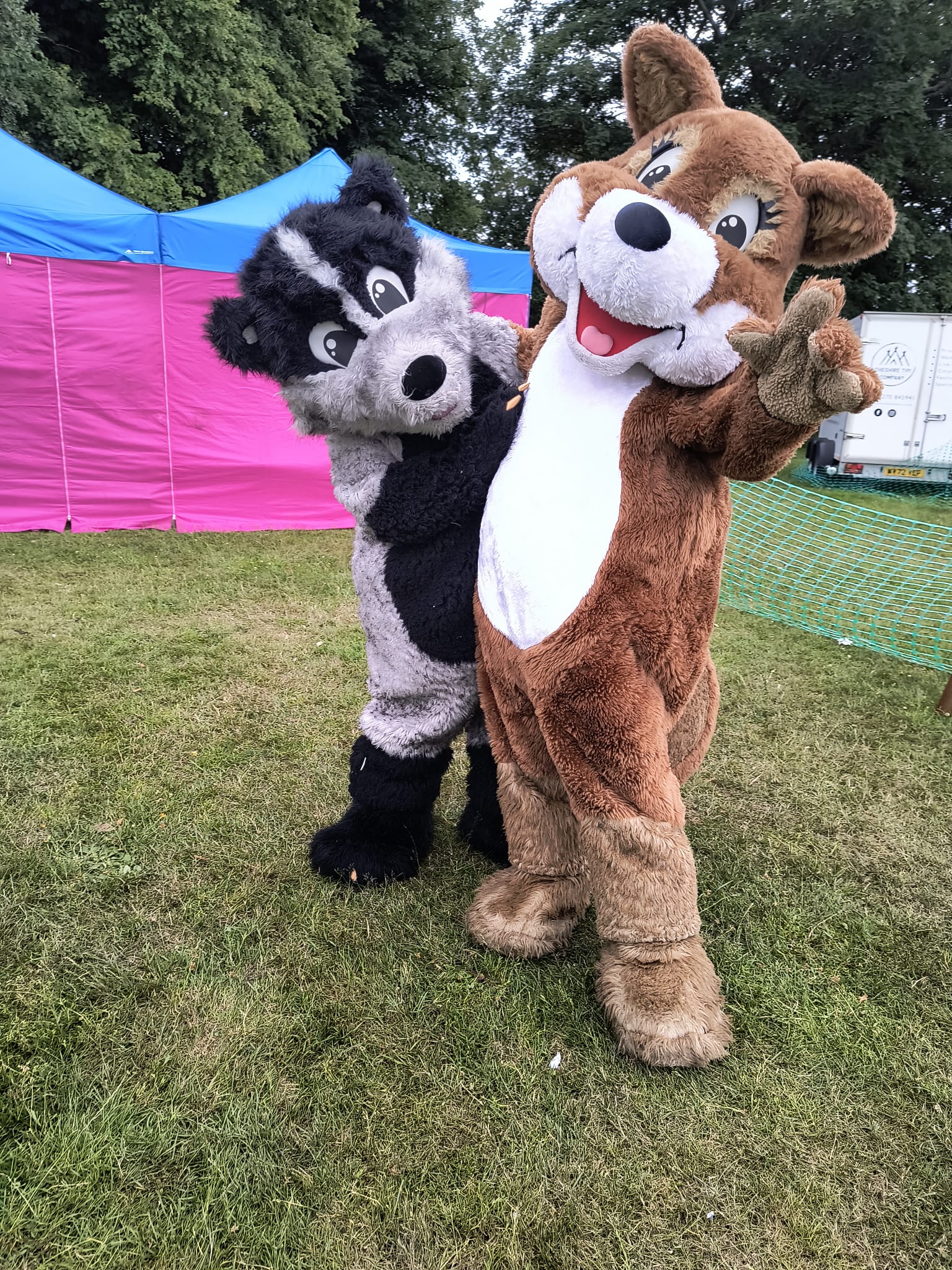 Bramble hugs Thistle as she points at the camera. They are stood on grass with a pink and blue gazebo behind them.