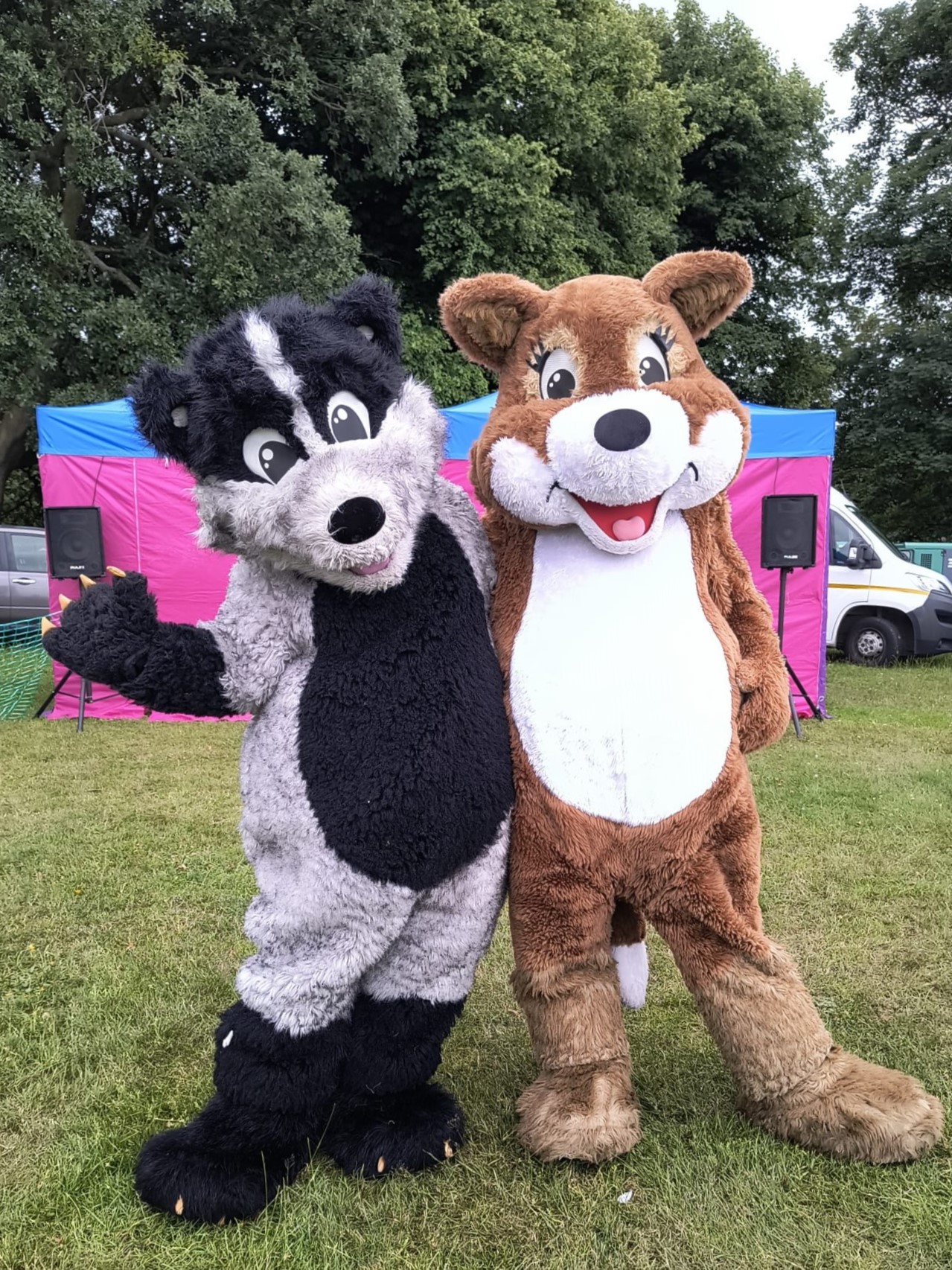 Bramble and Thistle stand side by side, waving at the camera.They are stood on grass with a pink and blue gazebo behind them.
