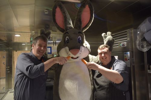 Truffles in the kitchen of a ship, posing with two chefs who are holding out carrots to him.