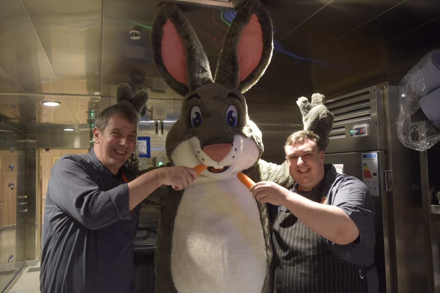 Truffles in the kitchen of a ship, posing with two chefs who are holding out carrots to him.