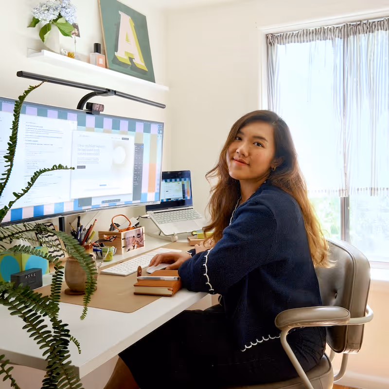 Woman with long hair seated at a desk in a bright home office, smiling at the camera with a large monitor and laptop in front of her.