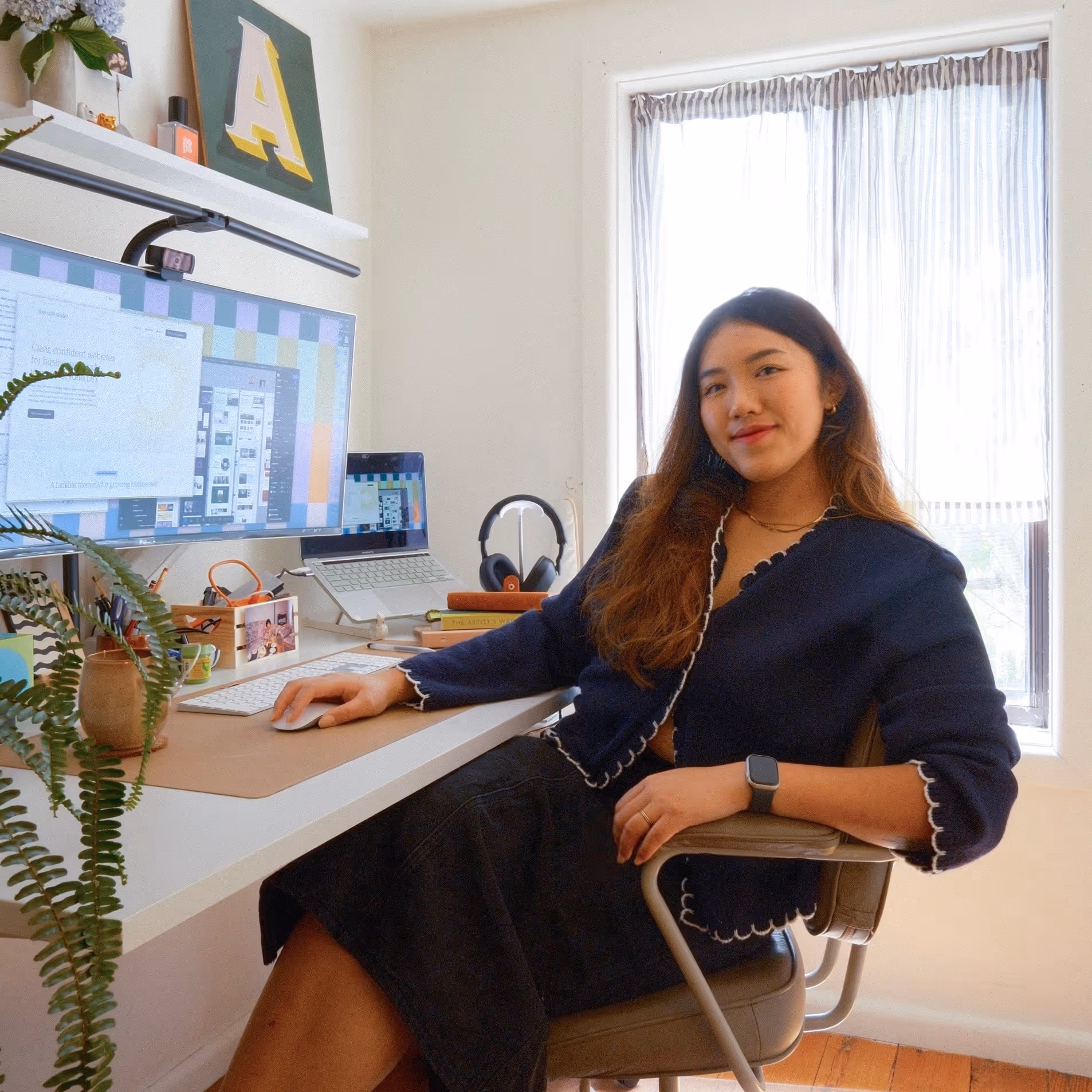 Woman sitting at a desk with computer monitors, smiling towards the camera in a bright home office.
