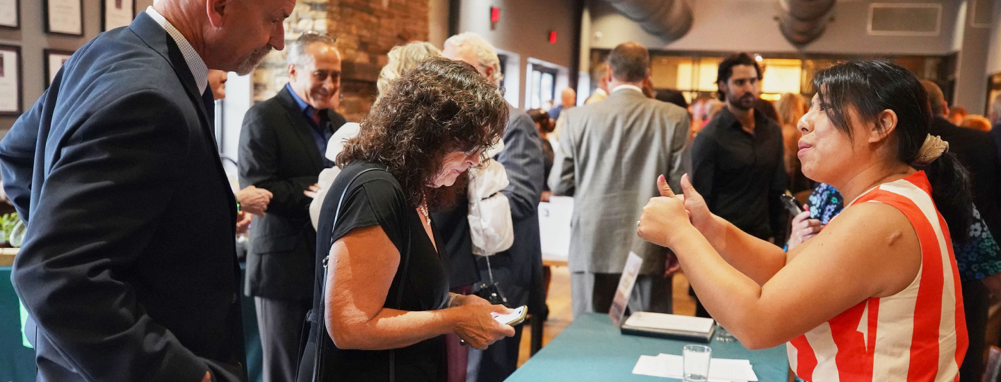 Woman in red and white striped sleeveless top giving a thumbs-up while talking to a woman holding a phone at an indoor event with multiple people in the background.