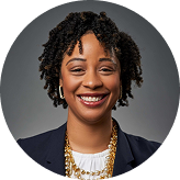 Smiling woman with curly hair wearing a navy blazer, layered necklaces, and hoop earrings against a gray background.