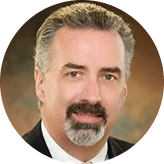 Portrait of a middle-aged man with short gray and dark hair, a mustache and goatee, wearing a dark suit and white shirt.