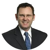 Smiling man wearing glasses, a dark suit, white shirt, and blue patterned tie against a white background.