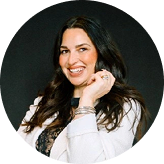 Smiling woman with long dark hair wearing a white jacket and silver jewelry against a dark background.