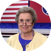Smiling elderly woman with gray hair wearing glasses, a yellow cardigan, and a blue top, with an American flag background.