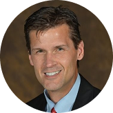 Portrait of a smiling man with short brown hair wearing a black suit jacket, light blue shirt, and red tie against a brown background.