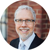 Smiling middle-aged man with gray hair, glasses, and beard wearing a suit and tie in front of a brick wall.