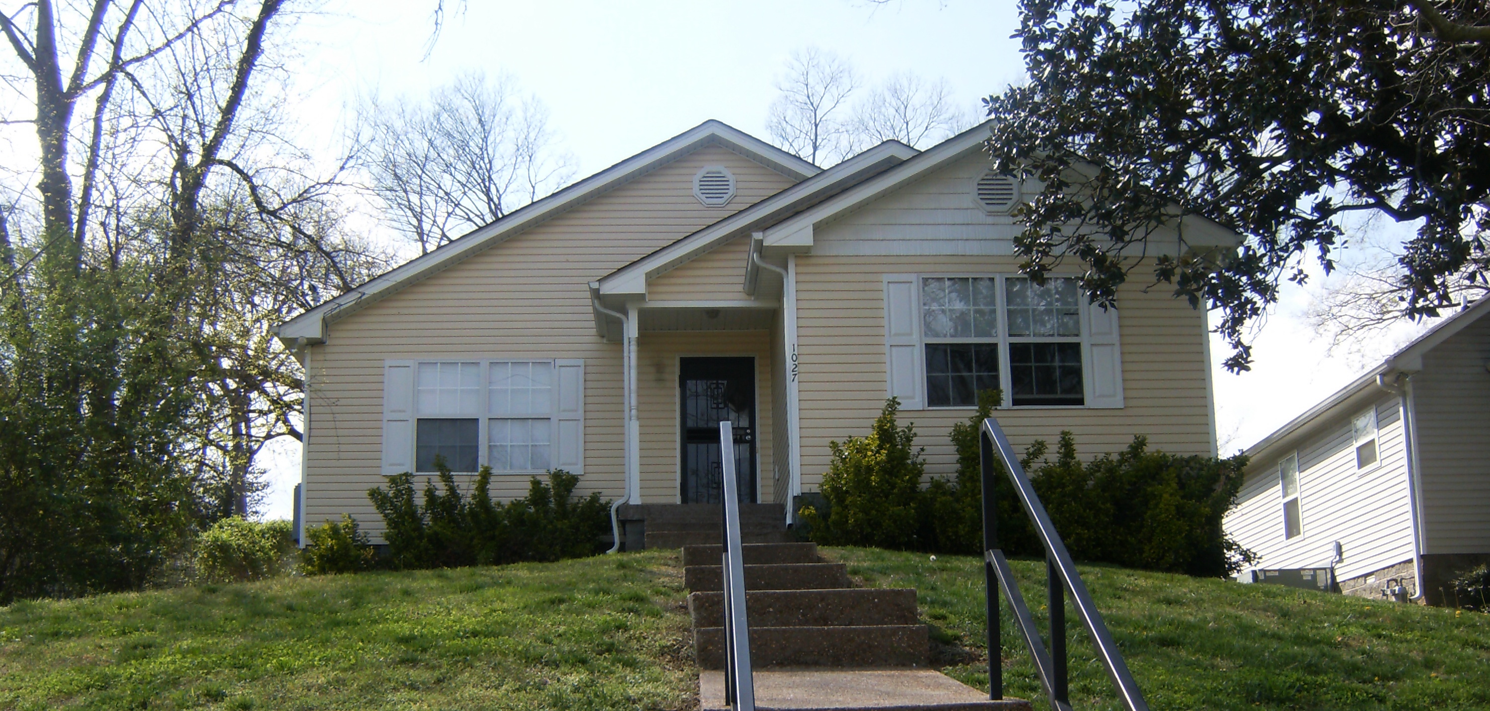 Light yellow single-story house with white trim, front stairs with black railings, and shrubs in front yard.