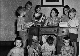 Black and white photo of a woman sitting at a desk with seven boys, some standing and some sitting on the floor, reading books.