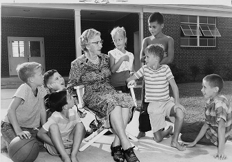 Black and white photo of an elderly woman sitting in a chair surrounded by seven young boys outdoors in a schoolyard or playground.